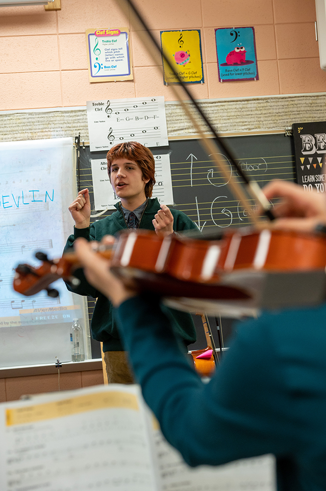 A violin in the foreground-only the players arms and left shoulder are seen. In the background a teacher stand talking in front of a blackboard