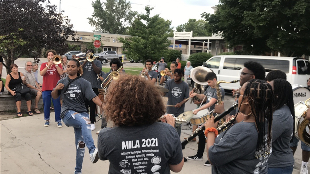 A group of students with various instrument stand in a circle outside while one person in the middle dances
